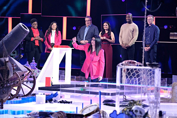 The host of Domino Masters, Eric Stonestreet (third from left), shouts “it’s topple time!” as Farah Bajwa (front) prepares to set off the first domino in front of judges. 