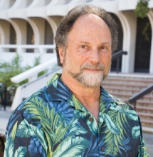 Professor Blumberg is standing in front of Langston Library at the UCI campus. He is wearing a dark blue button up shirt and is smiling softly.