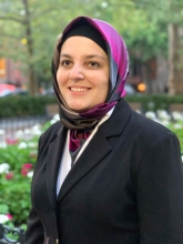 Gaze uplifted, Mona smiles.  Mona is wearing a black blazer over a white dress shirt, and a delicately wrapped purple and grey hijab.  Out of focus in her background is a vibrant green garden with white and pink flowers, framed by trees. 