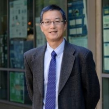 Professor Hu is standing in front of a building. He is wearing a gray blazer, a light blue dress shirt, and a blue tie.