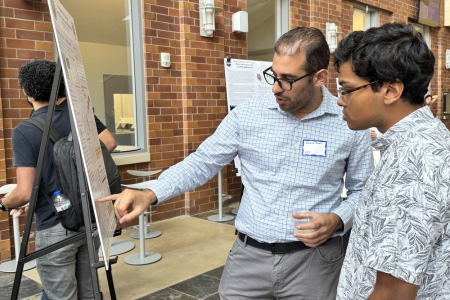 Two men standing near a poster, with one pointing at the poster.
